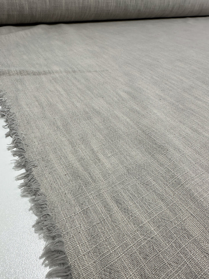 A close-up of a large roll of Super Cheap Fabrics' Basket Dyed Cotton in Dove, measuring 140cm, showcases its textured gray upholstery fabric with a subtle pattern and twill weave, complete with tassels along the edge.