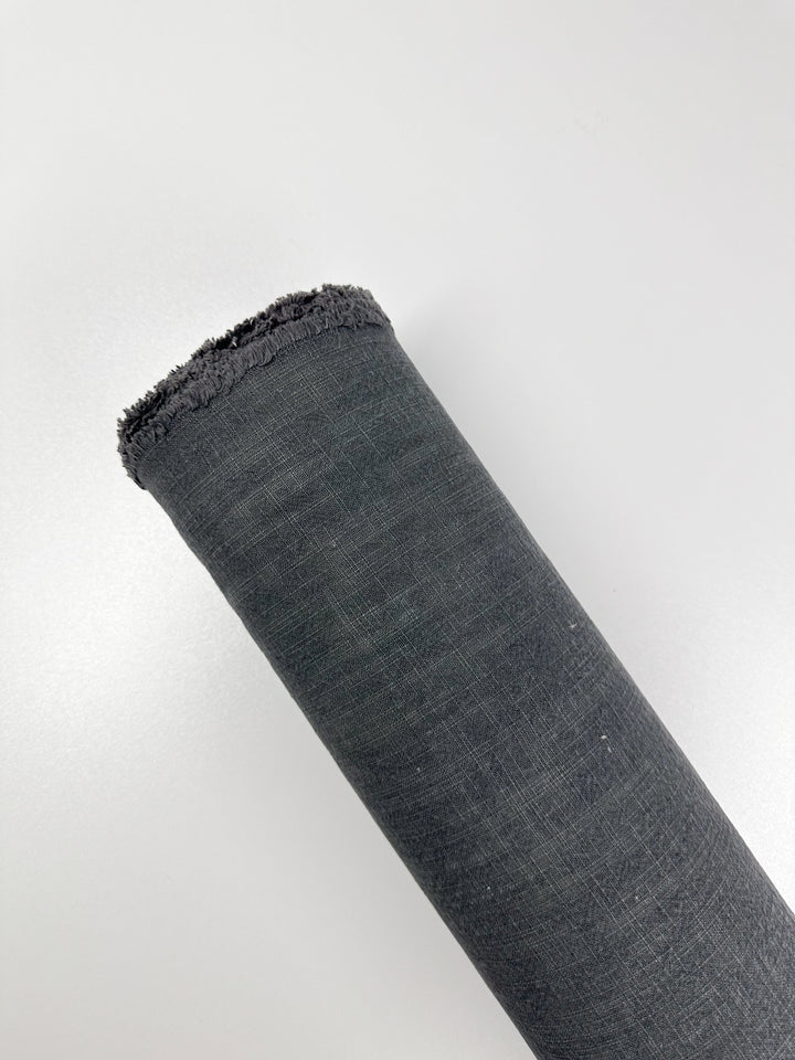 The Basket Dyed Cotton - Charcoal fabric roll, measuring 140cm and manufactured by Super Cheap Fabrics, is placed diagonally against a light gray background. This cylindrical textile is crafted from 100% cotton and displays a textured surface that highlights its twill weave.