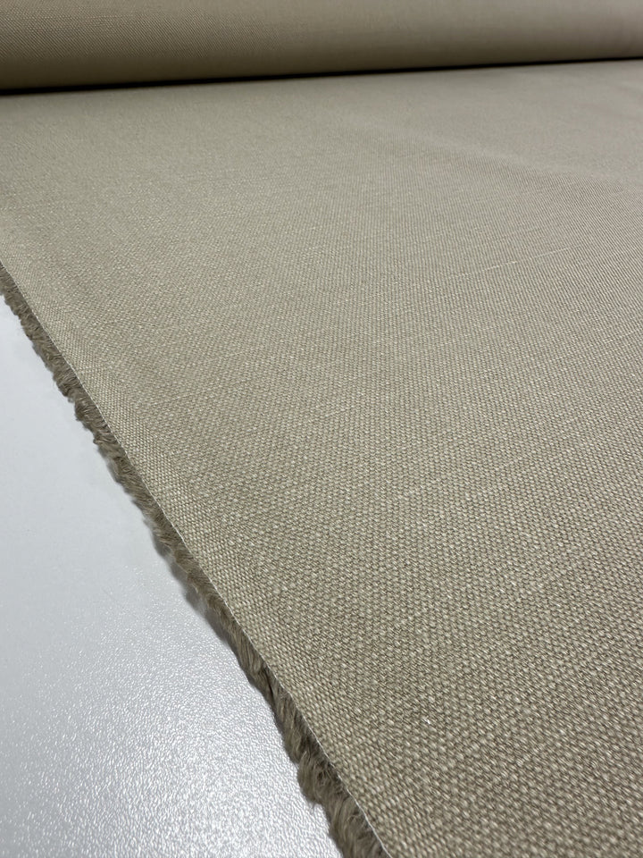 A close-up view of the Canvas Linen Denim - Peyote fabric from Super Cheap Fabrics reveals its heavyweight upholstery quality with a textured weave pattern in light beige. Partially unrolled, it showcases its clean edges and smooth surface on a white tabletop.