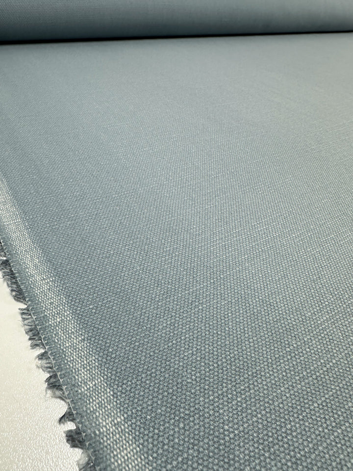 Close-up of a large roll of Canvas Linen Denim in the shade Blue Fog by Super Cheap Fabrics, featuring a subtle texture. The 140cm wide heavyweight fabric is spread out, covering the surface in the foreground, with visible fringed edges on the left side. The scene captures its smooth and even weave.