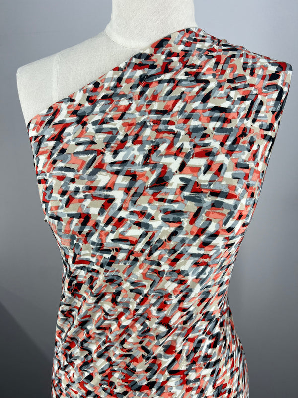 A mannequin is draped in Super Cheap Fabrics' Printed Lycra - Stroke - 150cm, a sleeveless polyester/spandex garment featuring an abstract pattern with scattered red, white, and black brush strokes. The medium-weight fabric appears slightly gathered at the sides. The background is a plain, neutral grey.