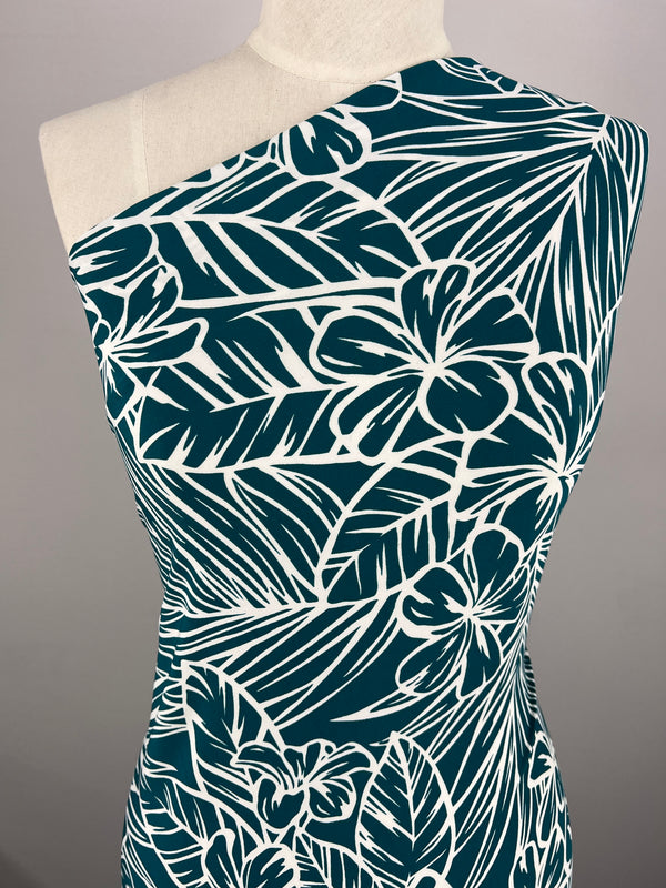 A dress form displays a one-shoulder garment adorned with an intricate tropical print. Designed using "Printed Lycra - Jasmint - 150cm" from Super Cheap Fabrics, the two-way stretch fabric showcases large white flowers and leaves set against a dark teal background. The dress form stands against a plain, neutral backdrop.