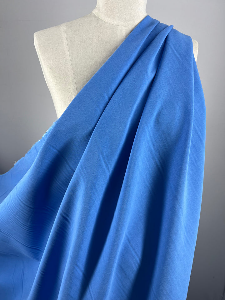 A close-up of medium weight suiting fabric from Super Cheap Fabrics draped over the shoulder of a white dress form, showcasing the texture and folds of the Silver Lake Blue - 145cm material. The dress form is positioned against a neutral gray background.