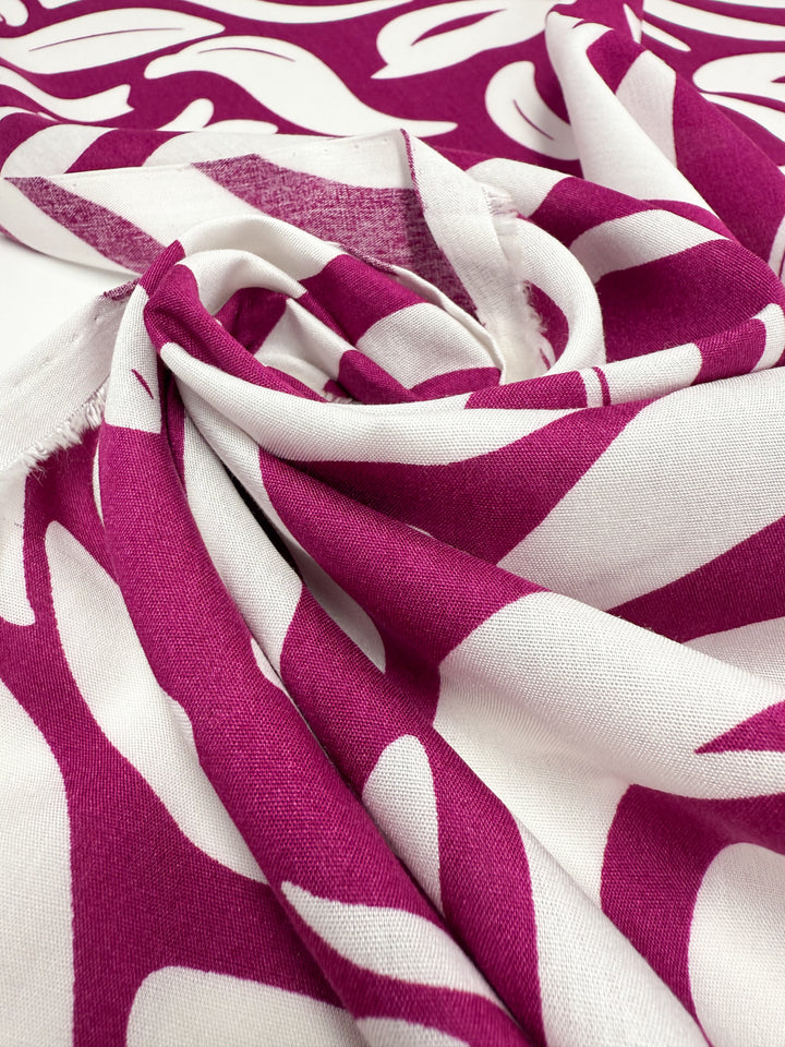 A close-up of Super Cheap Fabrics' Designer Rayon - Wild Bloom Fizz (145cm), featuring lightweight material with bold magenta and white abstract leaf prints, highlighting the vibrant colors, texture, and fabric folds.