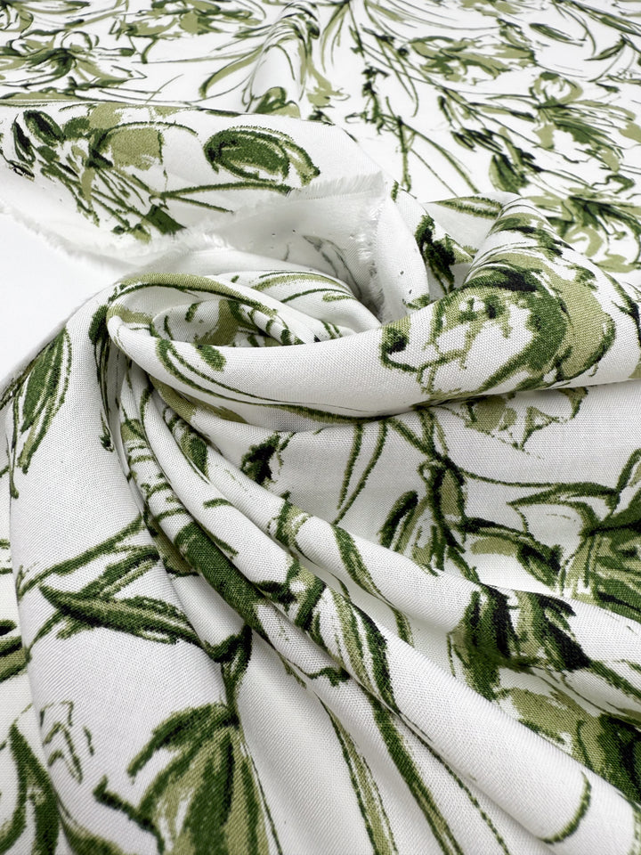 A close-up of Super Cheap Fabrics' Designer Rayon - Olive Sketch (145cm), showing white fabric with a green leafy vine pattern, loosely gathered and softly folded to highlight its texture and intricate botanical design.