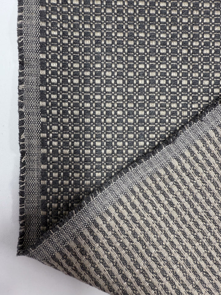 A close-up of Super Cheap Fabrics’ Upholstery Basketweave - Aria Steel (144cm) shows two layers with geometric patterns; one is folded to reveal the contrasting reverse. Slightly frayed edges highlight its premium quality.