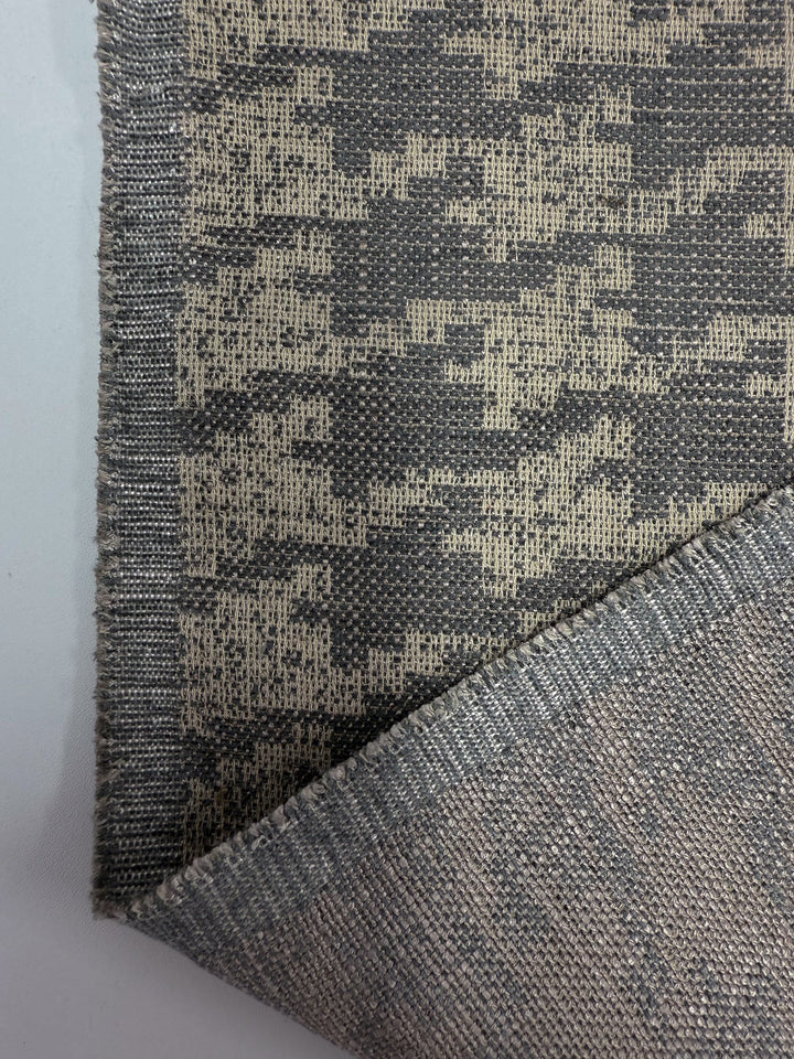 A close-up of Super Cheap Fabrics’ Upholstery Jacquard - Westwood Grey Dusk (154cm) shows its textured Australian weave, gray and tan abstract pattern, folded corner with reverse side visible, and detailed edge stitching.
