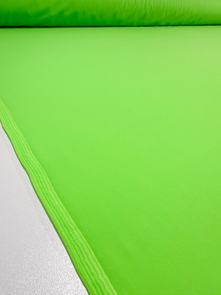 A close-up view of the Nylon Lycra - Green Flash - 160cm fabric roll by Super Cheap Fabrics, showcasing its vibrant green hue on a light-colored surface and emphasizing the texture and smoothness of its four-way stretch.