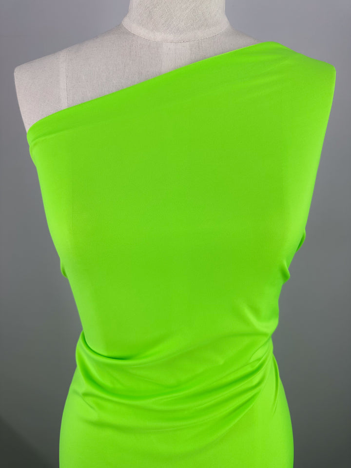 A mannequin showcases a sleek, modern one-shoulder dress made from the "Nylon Lycra - Green Flash - 160cm" fabric by Super Cheap Fabrics. This vibrant neon green garment, crafted with four-way stretch fabric, features a form-fitting silhouette and ruched detailing at the waist. The plain background accentuates the dress's striking color.