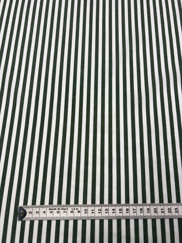 The Cotton Lawn - Fir - 150cm by Super Cheap Fabrics is displayed with alternating vertical green and white stripes. A measuring tape, marked in both inches and centimeters, is laid flat at the bottom edge to indicate the width of the stripes. This 100% cotton material features a silky finish.