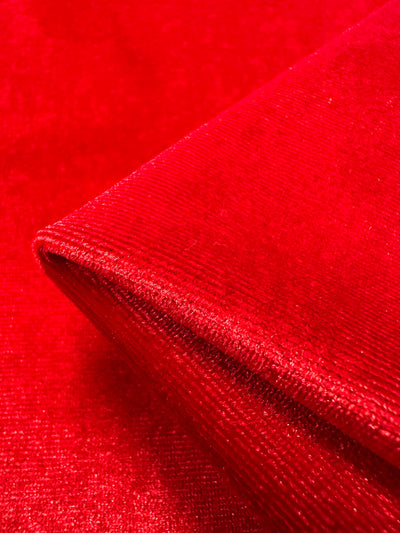 A close-up of the folded, shiny Mars Red fabric from Super Cheap Fabrics shows the medium-weight stretch velour's smooth, velvet-like texture and vibrant color that catches the light. Product: Stretch Velour - Mars Red - 160cm.