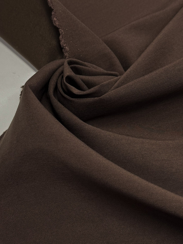 A close-up of Super Cheap Fabrics' Cotton Bengaline - Espresso Brown (145cm) displays its smooth texture, unfinished edges, and flexible stretch as the fabric is arranged in gentle folds and swirls.