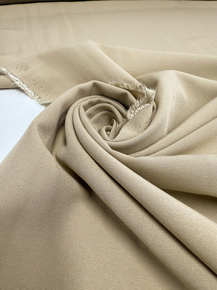 A close-up of Super Cheap Fabrics’ Cotton Bengaline - Sand Drift (150cm) shows the smooth, soft texture and gentle stretch of the fabric, laid flat with a central spiral to highlight its beige color and weave.