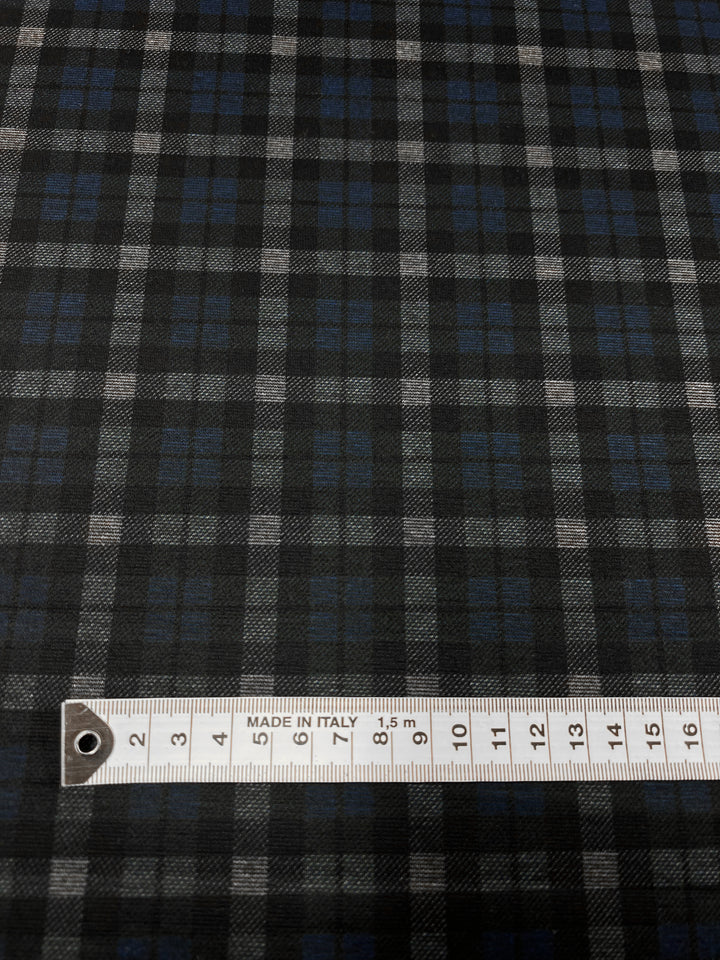 A close-up image of Super Cheap Fabrics' Ponte - Checkers - 150cm, showcasing a medium weight ponte fabric with a dark plaid pattern in black, white, and blue. A metal measuring tape featuring the inscription "MADE IN ITALY" is placed horizontally at the bottom of the image and measures up to 15 centimeters, ideal for crafting stylish pants.