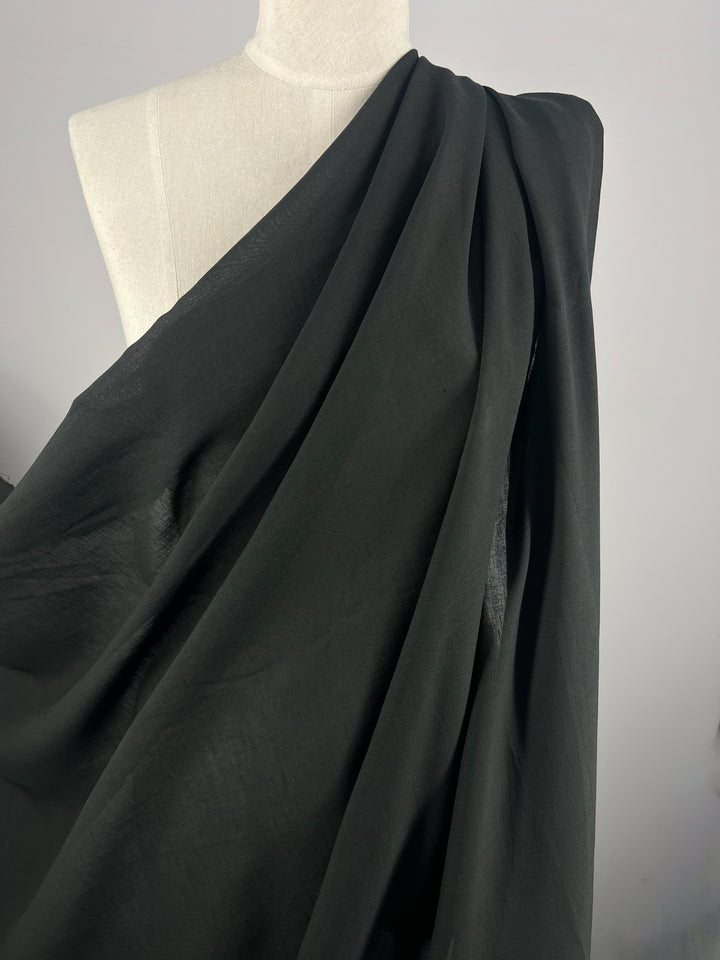 A white mannequin is draped with a large piece of Linen Voile - Bristol Black - 146 cm from Super Cheap Fabrics, which covers one shoulder and gathers elegantly over the torso. The background is plain and light-colored, focusing attention on the fabric's texture and flow on the mannequin.