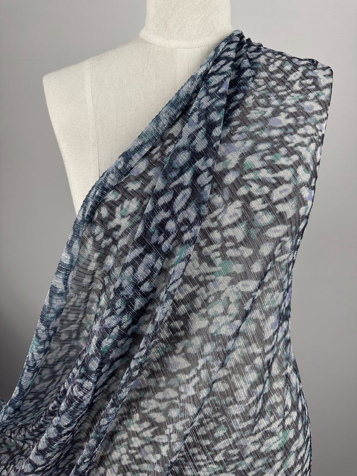 A mannequin is adorned with the Metallix Pleat - Ocean Leopard fabric from Super Cheap Fabrics, featuring a sheer, lightweight polyester material in an abstract black, navy, and white pattern that creates a textured, flowing appearance against a neutral gray background.