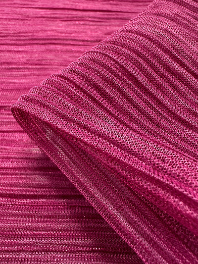 A close-up of the vibrant, hot pink Metallix Pleat fabric from Super Cheap Fabrics reveals its textured polyester with horizontal ridges. The lightweight 140cm-wide material is folded in the foreground, showcasing its intricate weave and shimmering metallic appearance.