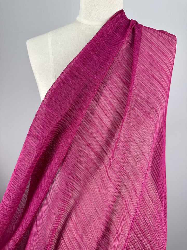 A mannequin is adorned with Metallix Pleat fabric from Super Cheap Fabrics, showcasing its sheer and textured hot pink material with metallic undertones. The lightweight fabric gracefully drapes with delicate crinkled lines over the shoulder, set against a plain light gray background.