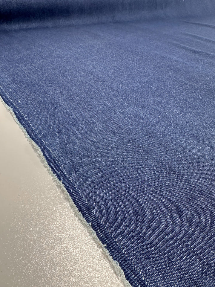 Close-up of a roll of Denim - Oceana - 150cm by Super Cheap Fabrics, featuring a medium weight blue denim fabric with a visible selvage edge, lying flat on a light-colored surface. The 100% cotton texture highlights the weave of the material.