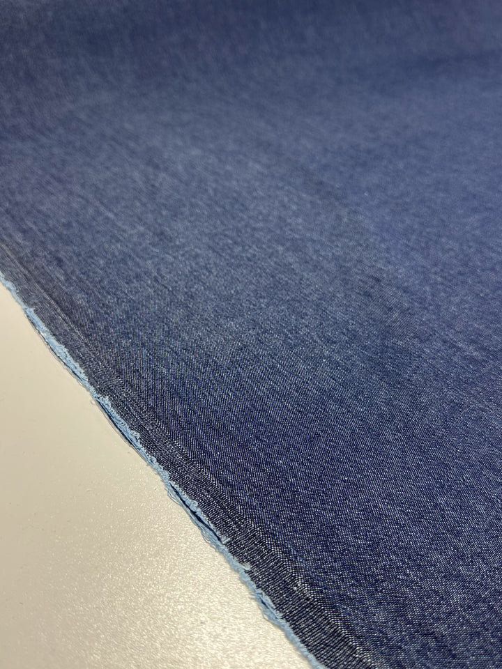 Close-up of the Super Cheap Fabrics Denim - Outer Space - 150cm, featuring a dark blue hue crafted from durable ring-spun yarns, with a light blue frayed edge, laid flat on a smooth white surface.