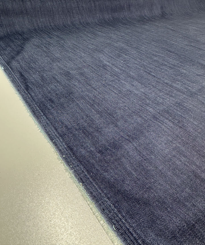 A close-up of a flat piece of Super Cheap Fabrics' Stretch Denim in Black Iris, measuring 140cm wide, is laid on a light surface. It showcases the texture and raw edges of the fabric, which is made from ring-spun cotton and appears smooth with a slight reflective quality under the lighting.