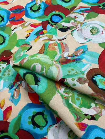 A close-up view of the “Designer Rayon - Eloise - 145cm” fabric by Super Cheap Fabrics showcases its vibrant, abstract floral design in red, green, blue, and white against a beige background. The fabric is gently folded at the center of the image.