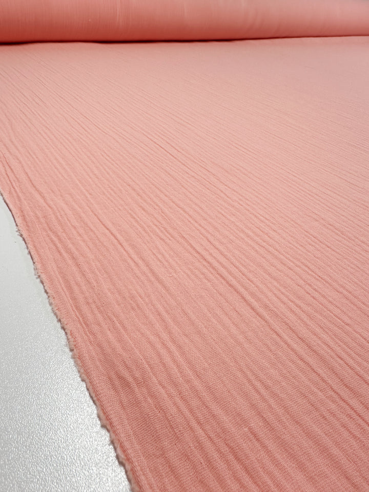 A close-up of a large roll of Muslin - Peach Parfait - 135cm by Super Cheap Fabrics, featuring a ribbed texture and spread out on a flat surface. The organic fabric appears soft and showcases a gentle sheen.