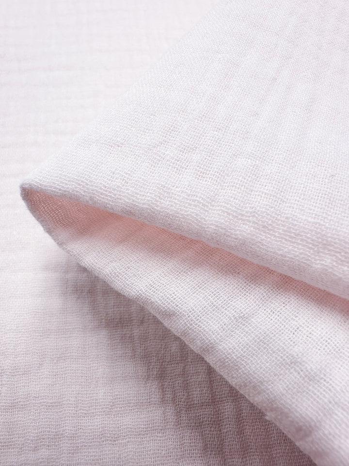 A close-up view showcases the Muslin - Slightly Pink - 135cm from Super Cheap Fabrics. The fabric features a soft, pale pink hue with a subtly textured, grid-like pattern. When folded, the reusable cloth highlights its lightweight and airy quality, and the visible texture suggests a gentle and delicate material.