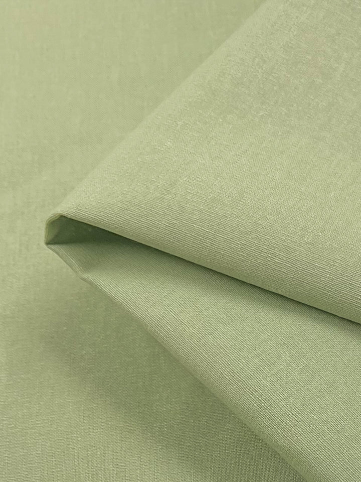 A close-up view of a folded piece of Super Cheap Fabrics' Linen Blend - Swamp - 147cm, displaying its light green hue and slightly textured, smooth surface. The fabric edges are neatly aligned, highlighting the quality and evenness of the material, reminiscent of the delicate beauty of a Four Leaf Clover.