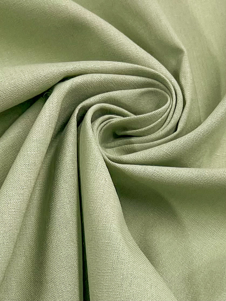 A close-up view of the Linen Blend - Swamp by Super Cheap Fabrics, showcasing its light green hue and slightly coarse texture, gathered and twisted into a spiral pattern. The light to medium weight fabric features visible weave details and a matte finish.