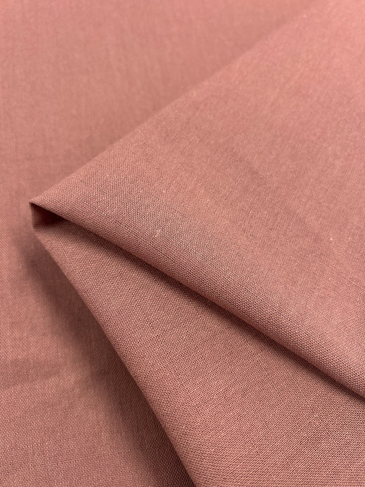 A close-up image of the Super Cheap Fabrics' Linen Blend in Muted Clay, which is 145cm wide. The linen blend fabric displays a smooth texture with slight folds, demonstrating its light weight as it gently overlaps and creates soft creases. The consistent color throughout makes it an excellent choice for both clothing and upholstery projects.