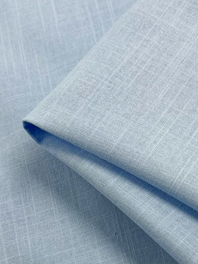 A close-up of the Linen Blend - Dutch Canal fabric from Super Cheap Fabrics, featuring a light blue hue with a subtle grid pattern. This neatly folded, 147cm wide linen cotton blend showcases its lightweight texture and intricate weave details. The smooth surface and soft color suggest it could be ideal for garments or home textiles.