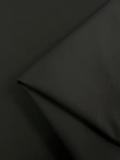 Close-up of the Designer Suiting - Noir - 145cm by Super Cheap Fabrics. The smooth, folded black fabric features a matte finish and slight texture, with a sharp fold creating a clean, angular line. This medium to heavy weight material is perfect for pants and dresses, with the background entirely made up of the same fabric for uniformity.