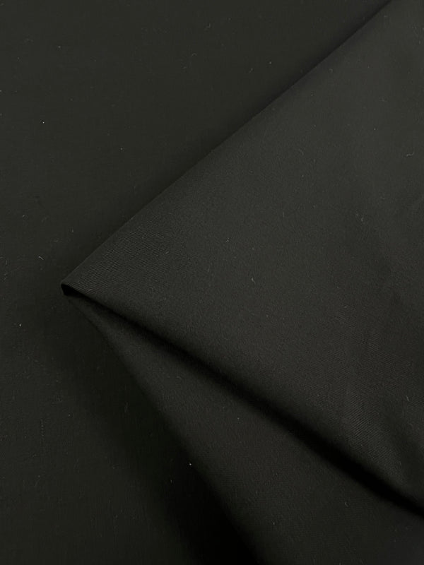 Close-up of the Designer Suiting - Noir - 145cm by Super Cheap Fabrics. The smooth, folded black fabric features a matte finish and slight texture, with a sharp fold creating a clean, angular line. This medium to heavy weight material is perfect for pants and dresses, with the background entirely made up of the same fabric for uniformity.