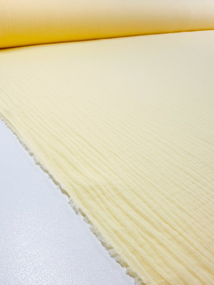 A close-up of a roll of Muslin - Pastel Yellow - 135cm by Super Cheap Fabrics reveals its delicate horizontal ridges. This organic fabric unfurls gracefully across a smooth, white surface, showcasing its textured beauty.