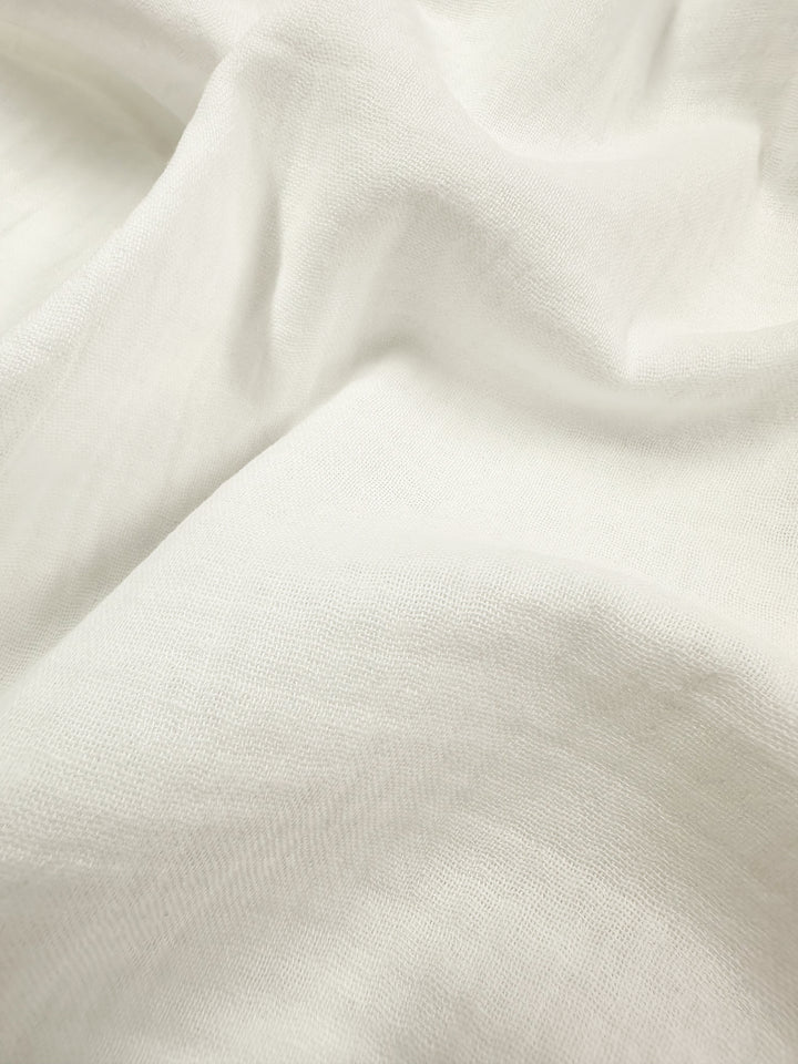 A close-up image of fabric branded as Super Cheap Fabrics' Muslin - Ivory - 135cm reveals its organic nature with a soft, textured surface. The material is gently folded, creating subtle shadows and highlights to showcase the delicate charm of this lightweight muslin fabric.