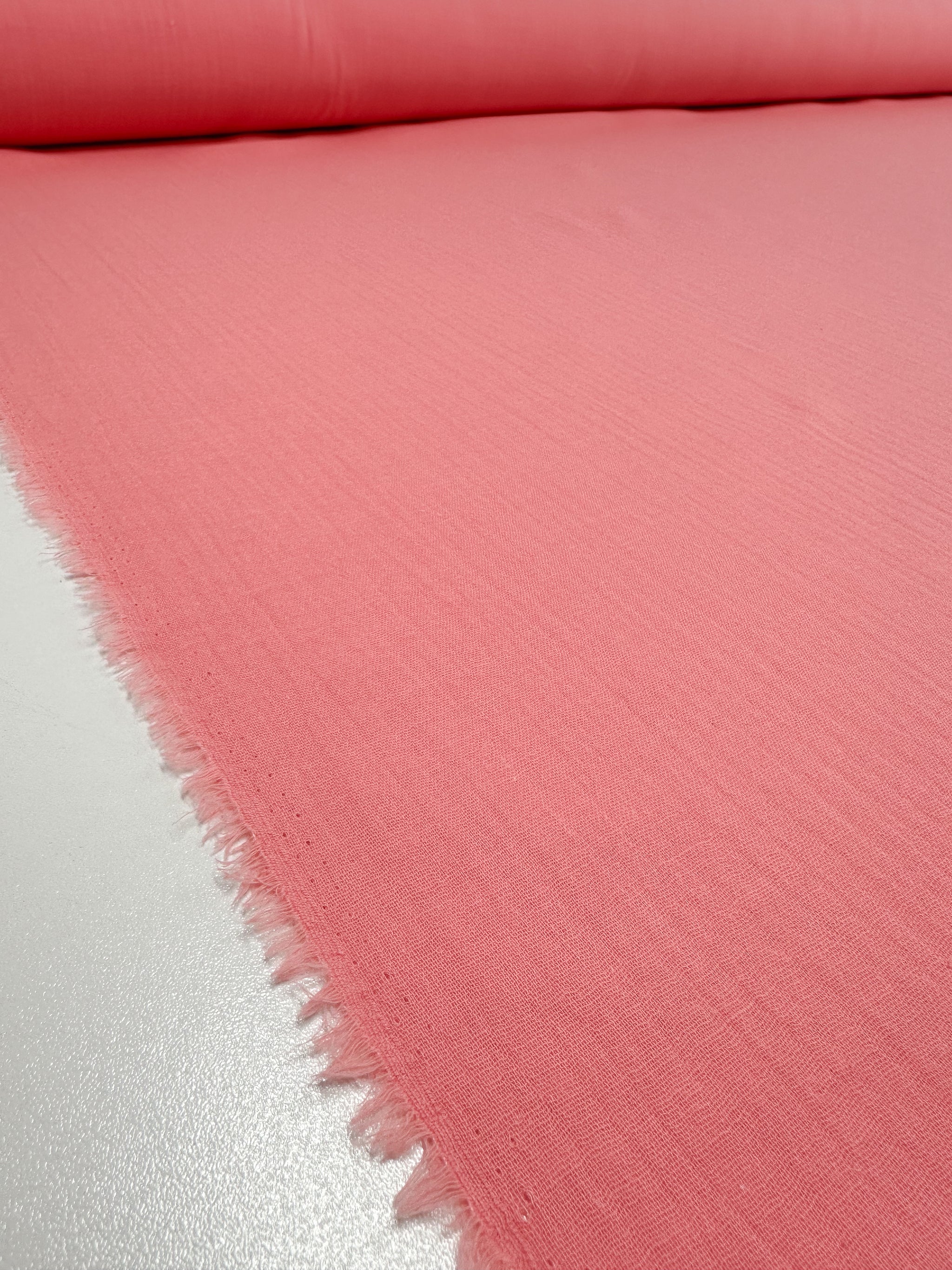 A close-up view of a roll of Super Cheap Fabrics' Muslin in Flamingo Pink, featuring a slightly textured surface and slightly frayed edges. This organic fabric roll is placed on a flat, light-colored surface.