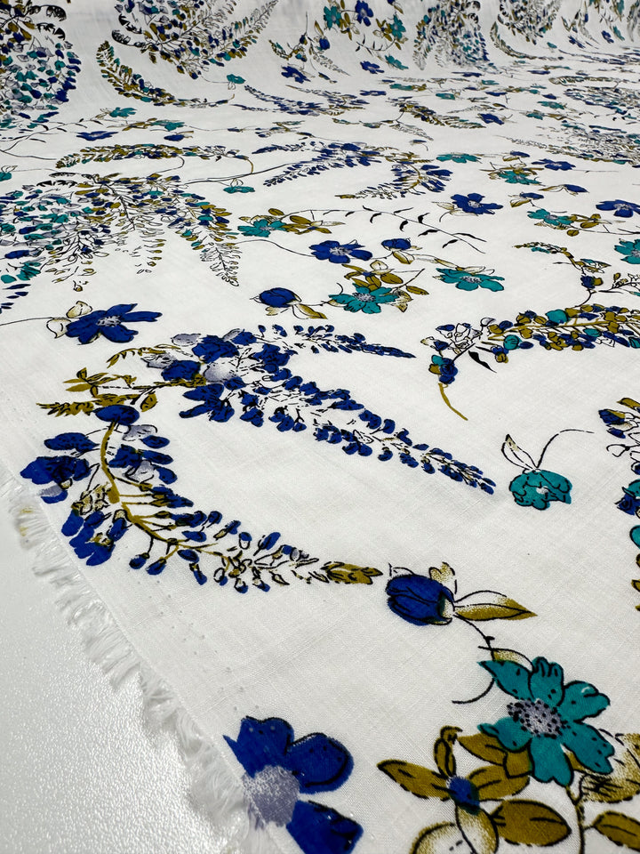 Presenting the Linen Blend - Royal Vine, courtesy of Super Cheap Fabrics: a close-up captures the exquisite breathable linen adorned with a floral pattern, featuring blue and teal blossoms intertwined with lush green leaves. This lightweight design elegantly embellishes the fabric's surface, making it an ideal choice for stylish summer attire. The fabric measures 145cm in width, offering ample material for your creative projects.