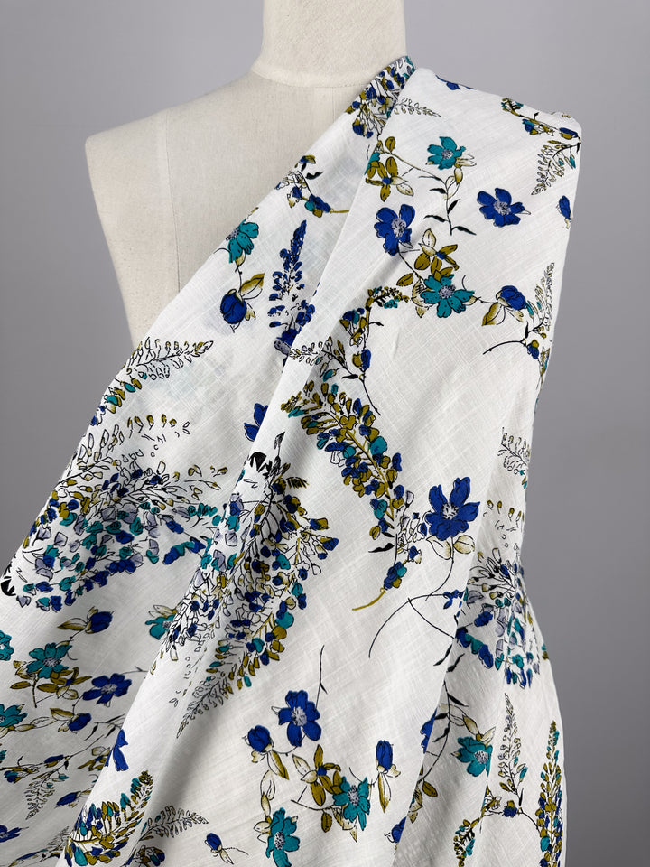 The Linen Blend - Royal Vine - 145cm from Super Cheap Fabrics is elegantly draped over a mannequin, featuring a floral pattern with blue and yellow flowers and green leaves. This breathable fabric showcases its texture and design beautifully, making it ideal for chic summer outfits.