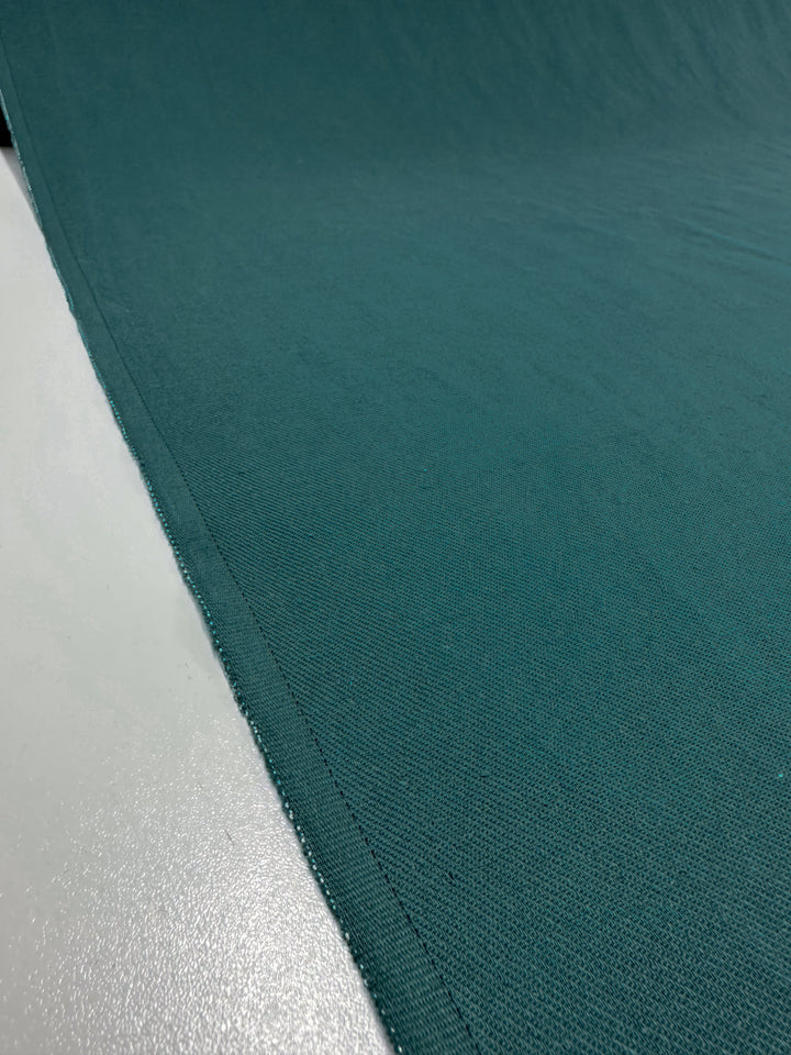 A close-up of Super Cheap Fabrics' Pure Washed Linen in Jasper, measuring 140cm, showcases the teal hue and visible stitching along the edge on a smooth white surface. The slightly coarse texture indicates its sturdy quality, making it suitable for a variety of textile applications.