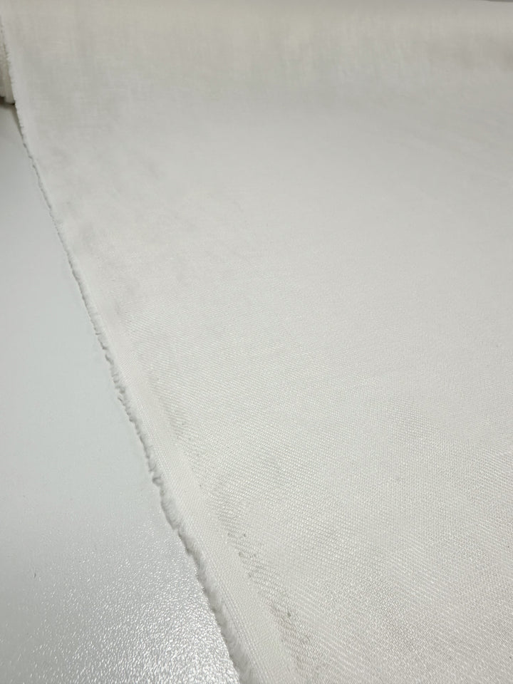 A close-up of a roll of Pure Washed Linen - Lucent White - 140cm by Super Cheap Fabrics, with its edge clearly visible as it rests on a smooth surface. The texture appears soft and slightly textured, indicating it's lightweight fabric ideal for clothing or upholstery.