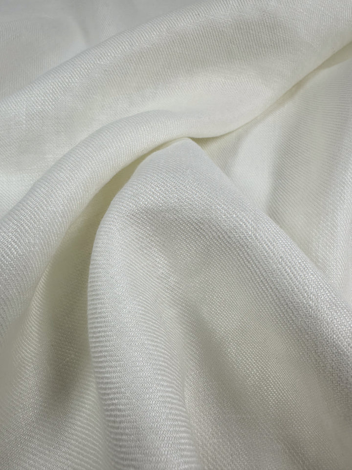 A close-up of the Pure Washed Linen in Lucent White by Super Cheap Fabrics reveals a lightweight and soft fabric with a smooth, slightly wavy texture. The material features a subtle sheen and an intricate weave, which creates gentle folds and highlights across its 140cm wide surface.