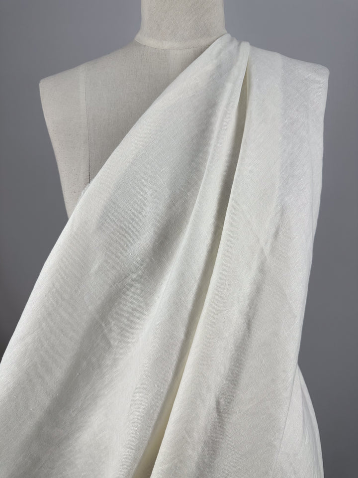 On a mannequin, the Pure Washed Linen - Lucent White from Super Cheap Fabrics is elegantly draped. The lightweight, textured fabric flows asymmetrically over one shoulder, creating a sophisticated and classical look. Against a plain gray backdrop, the subtle details of the linen are beautifully highlighted.