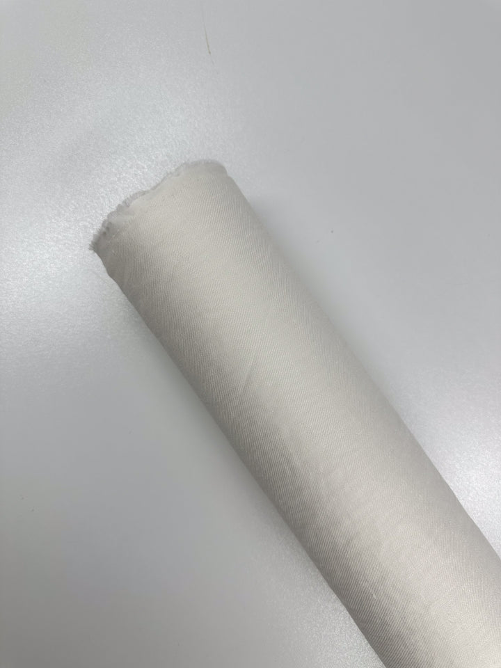 A roll of Super Cheap Fabrics' Pure Washed Linen in Lucent White, measuring 140cm, rests on a light grey surface. The fabric features a smooth, unpatterned texture with a slightly frayed edge at the top, highlighting its lightweight elegance.