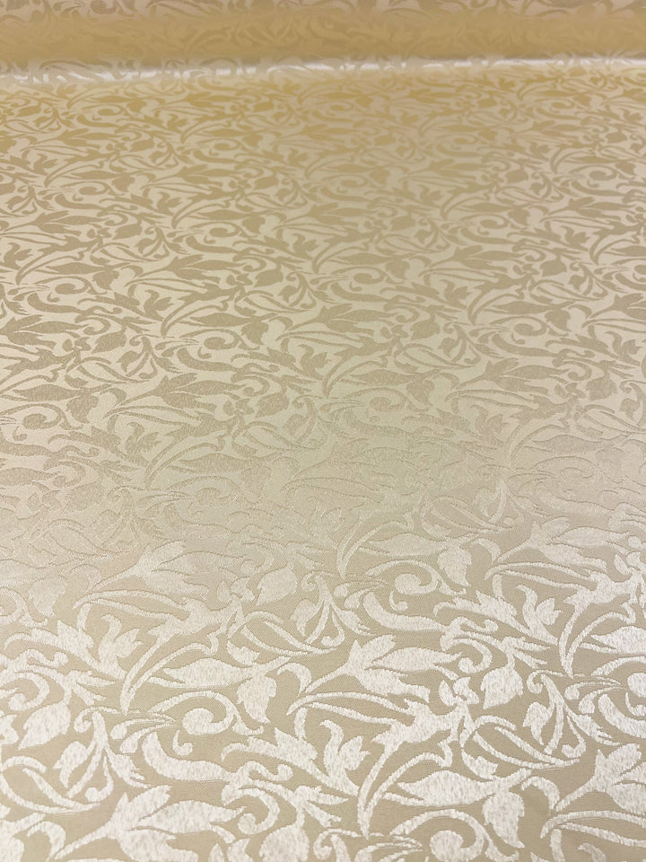 A close-up of the Designer Damask fabric from Super Cheap Fabrics in the Rutabaga color reveals its ornate, floral pattern. The intricate, swirling leaf motifs in a slightly darker shade create a subtle contrast that highlights the elegance of this luxurious, beige fabric measuring 155cm.