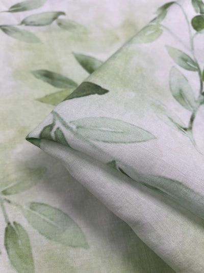 Close-up of the Designer Cotton - EUCAS - 150cm from Super Cheap Fabrics, a soft, folded lightweight cotton fabric with a light green and white leaf print pattern. The design features delicate, watercolor-style leaves over a muted, washed-out background, making it perfect for luxury sewing projects due to its gentle and calming appearance.