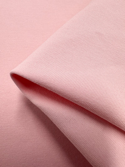 A close-up shows the Powder Pink Cotton Lycra by Super Cheap Fabrics, 167cm wide, folded diagonally. The medium-weight cotton has a ribbed texture and a breathable, smooth appearance that subtly highlights its fine textile details.