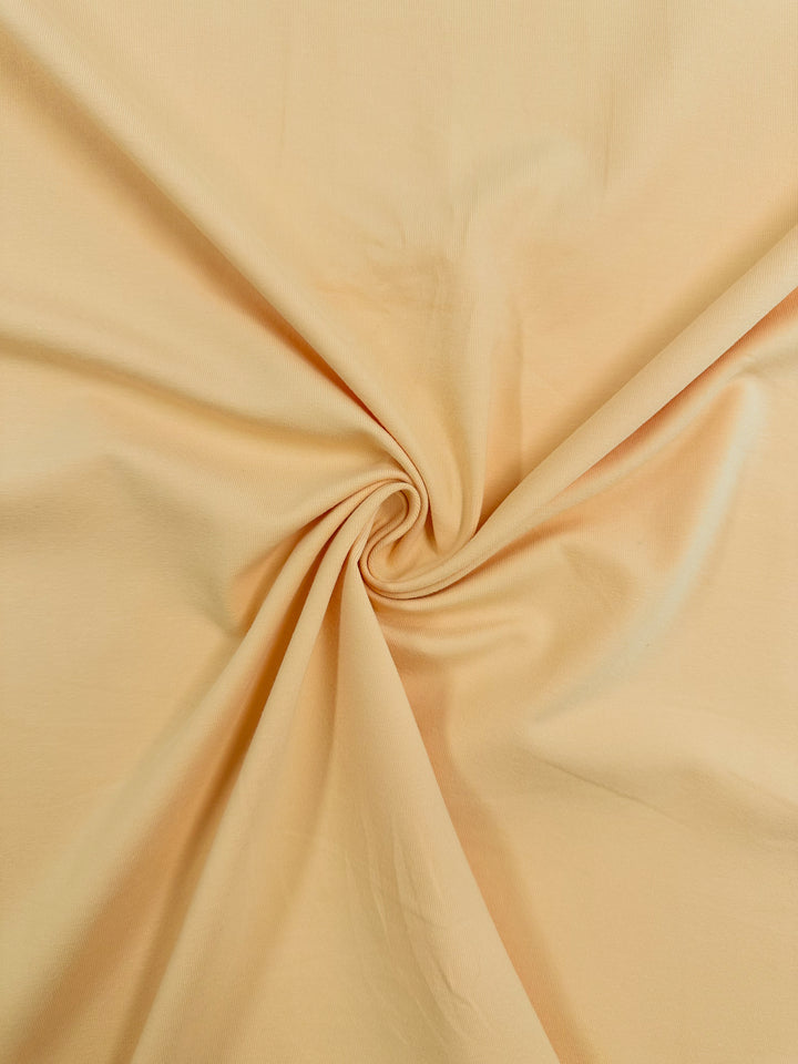 A close-up of Cotton Lycra - Caramel Cream by Super Cheap Fabrics reveals soft folds and texture with a central swirl pattern. The smooth, beige material is 165cm wide and drapes gently, showcasing its breathable qualities.