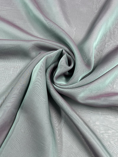 A close-up of the sheer Designer Shot Chiffon - Pearl Blue from Super Cheap Fabrics, beautifully arranged in a swirling pattern, highlights its soft green hue. The light accentuates subtle variations in this lightweight, 150cm fabric, lending it a delicate and airy appearance.