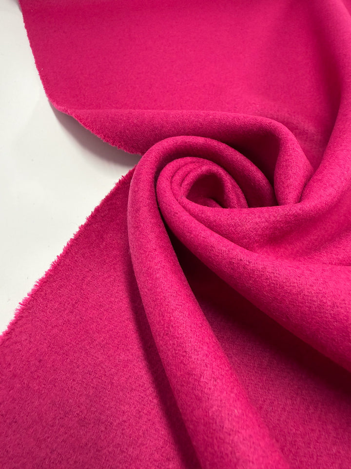 A close-up of Super Cheap Fabrics' Wool Cashmere - Vivacious (148cm) in bright pink, featuring its premium soft texture with a smooth surface and one frayed edge, partially rolled and twisted on a white background.